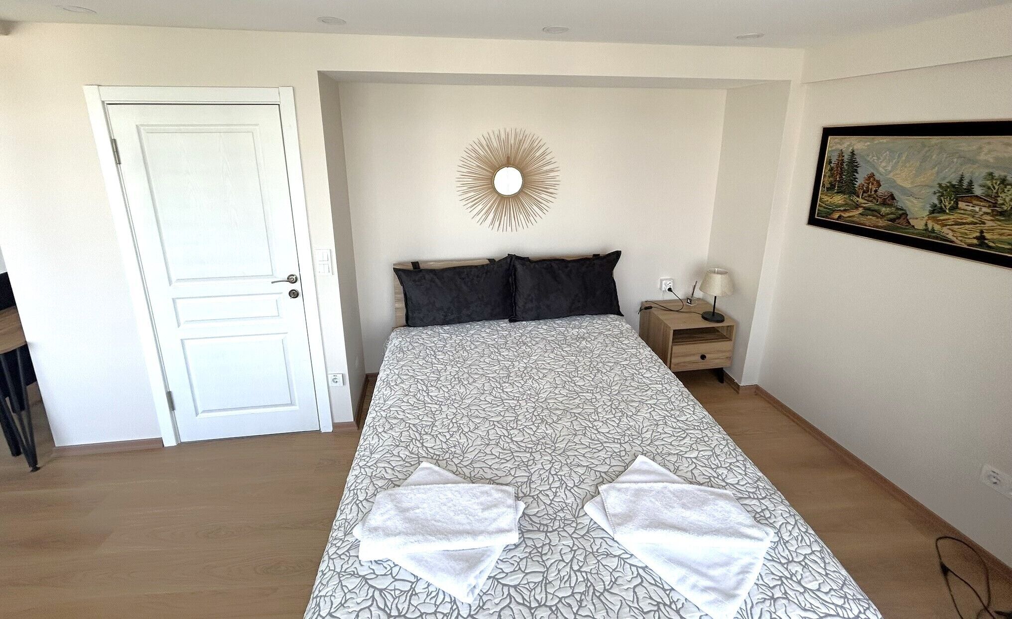 Een slaapkamer met een bed, nachtkastje, een spiegel aan de muur en een schilderij.