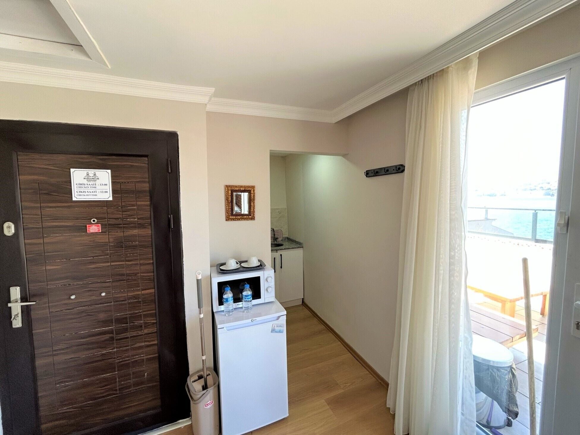 A hotel room with a wooden door, a small kitchenette, a microwave, and a view of the balcony.