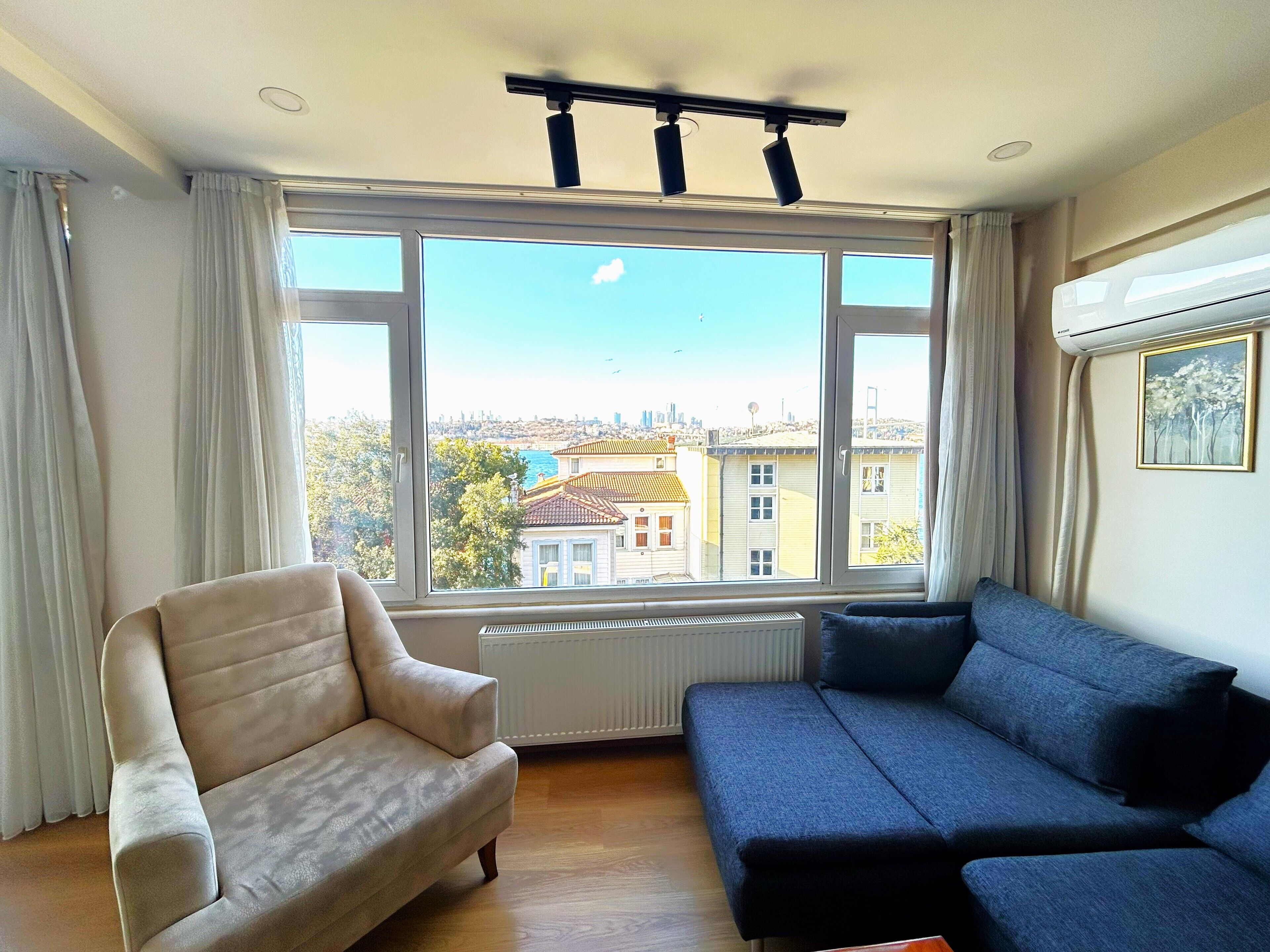 Een kamer met een groot raam, een beige fauteuil, een blauwe bank en een radiator.