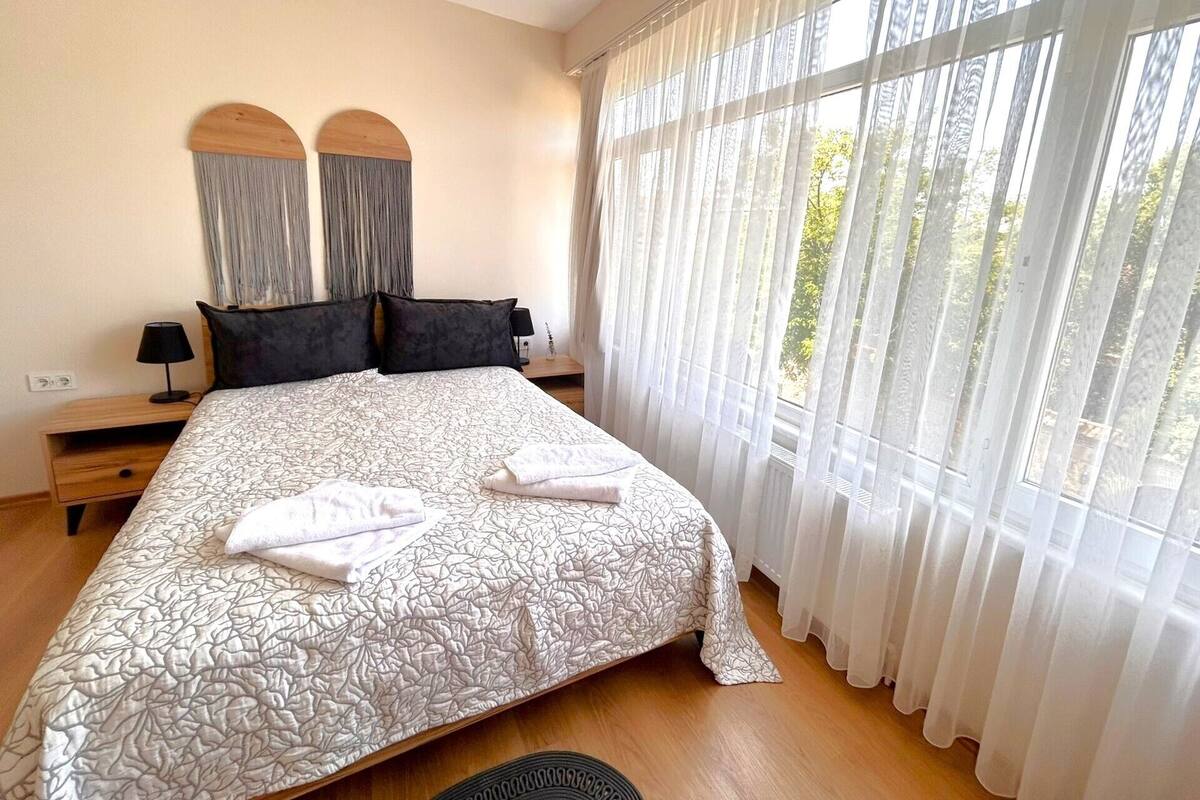 Un lit bien fait, recouvert d’une couvre-lit à motifs, avec deux oreillers et une serviette pliée. On y trouve une table de chevet avec une lampe et une étagère fixée au mur. La pièce est dotée de grandes fenêtres avec des rideaux légers, qui laissent entrer la lumière naturelle.