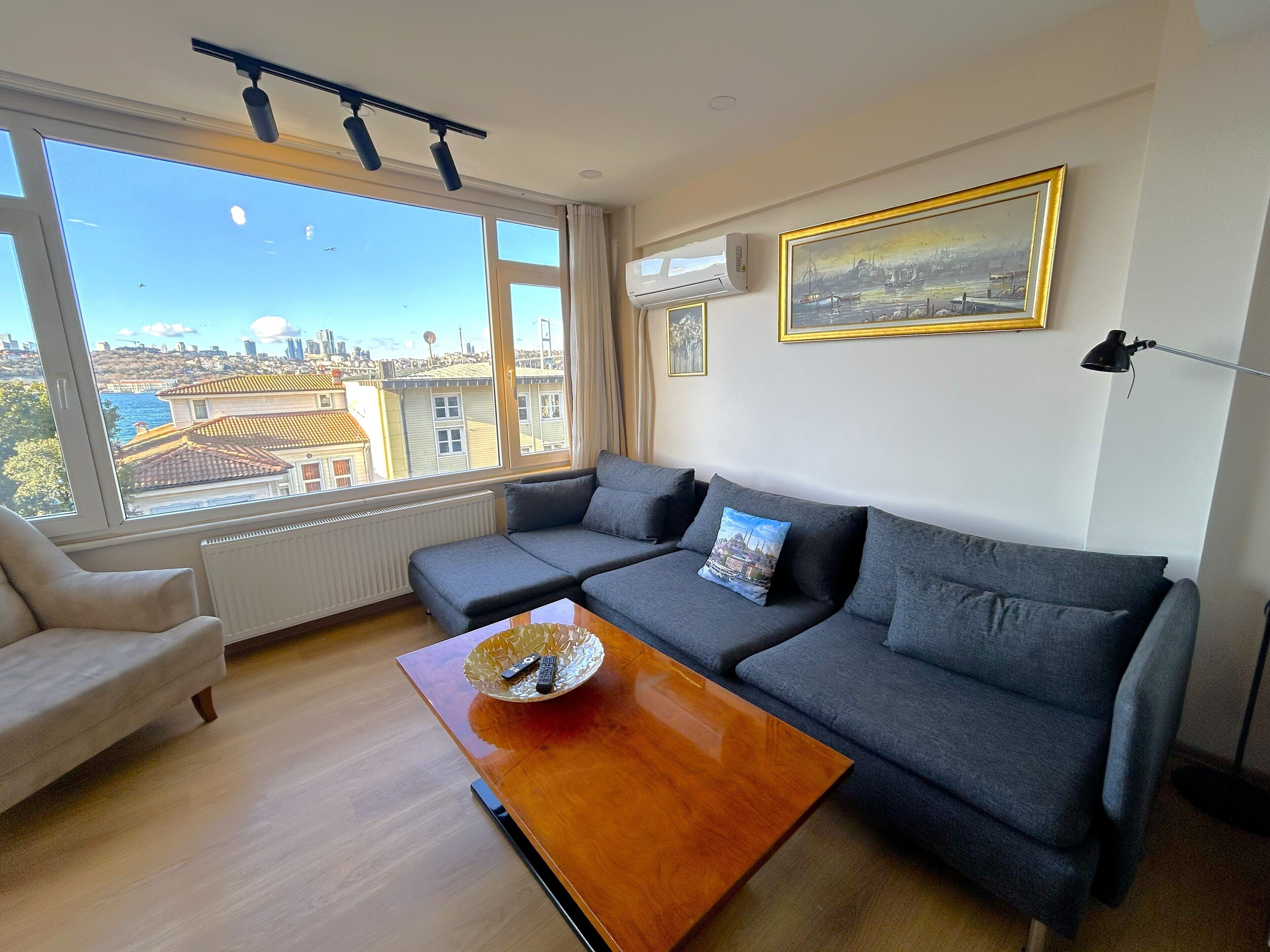 Un soggiorno moderno con un divano grigio, un tavolino in legno e vista sulla città attraverso ampie finestre.