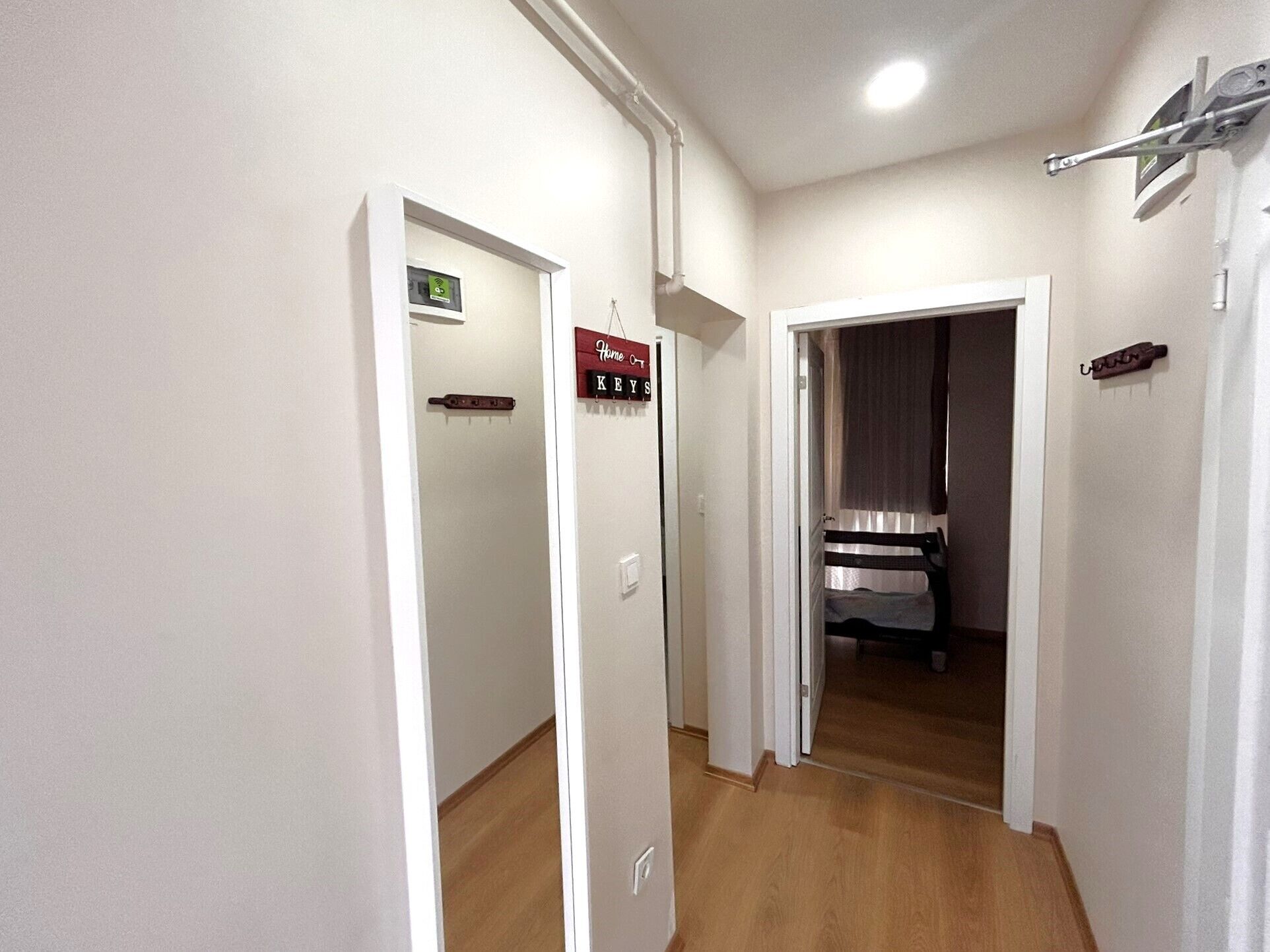 A hallway with a wooden floor, white walls, and a door leading to a room with a bed and a window.