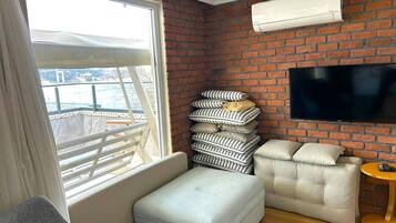 Habitación de hotel con pared de ladrillo, un televisor de pantalla plana sobre un sofá, una ventana con vista a un balcón y una pila de almohadas.