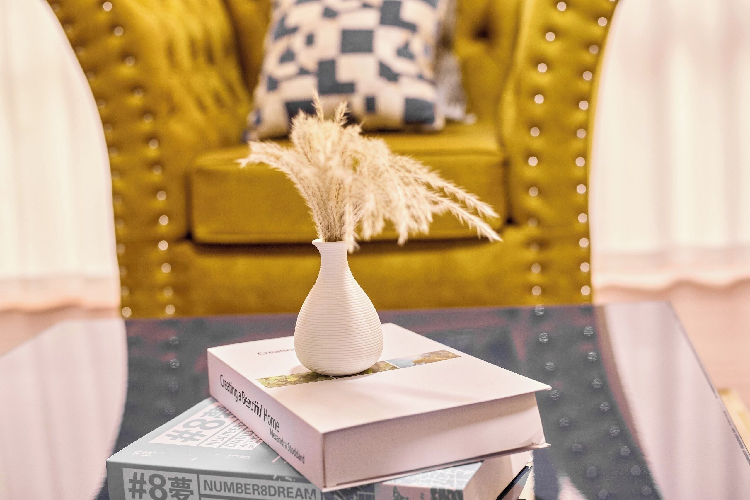 Un jarrón blanco con plumas de plumas de pampa seca sobre una mesa, junto a un libro titulado 