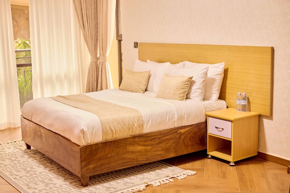 Ein ordentlich bezogenes Bett mit Holz-Kopfteil, ein Nachttisch mit Schublade und ein Fenster mit Vorhängen.