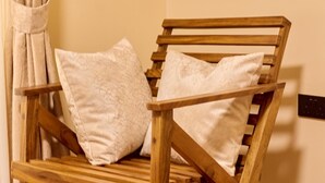 A wooden chair with two pillows, positioned against a wall with a curtain. - Sand Grains Residences 007 (Entebbe)