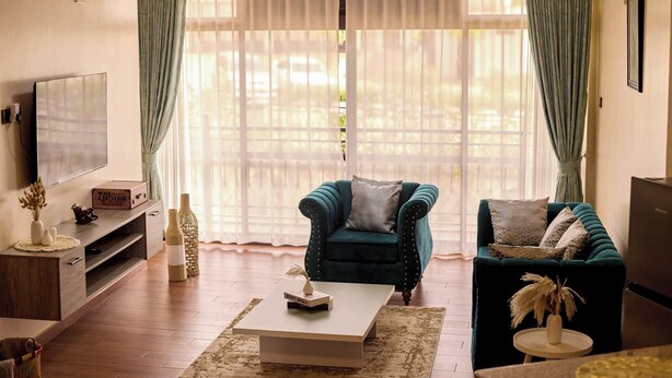 A living room with a teal armchair, a dark sofa, a coffee table, and a TV on a wooden stand. - Sand Grains Residences 007 (Entebbe)