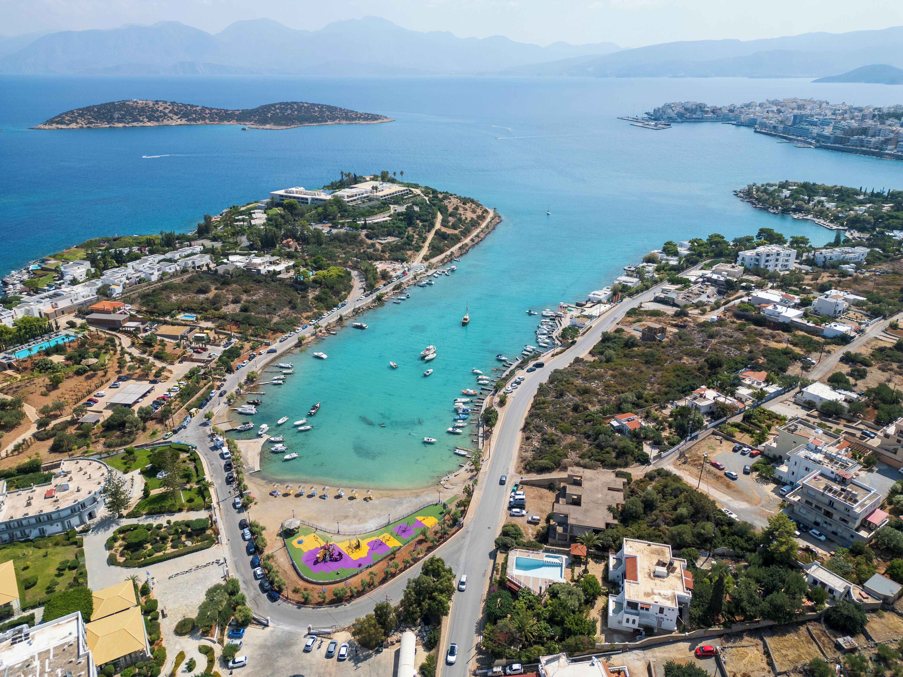 An aerial view of a coastal area with a marina, buildings, and a road.