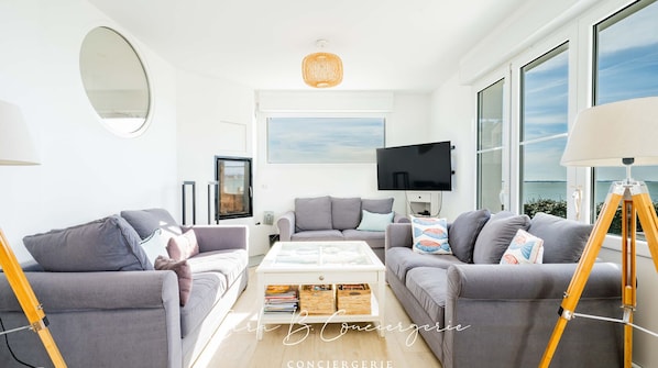 A modern living room with two grey sofas, a coffee table, and a large window offering a view of the sea.
