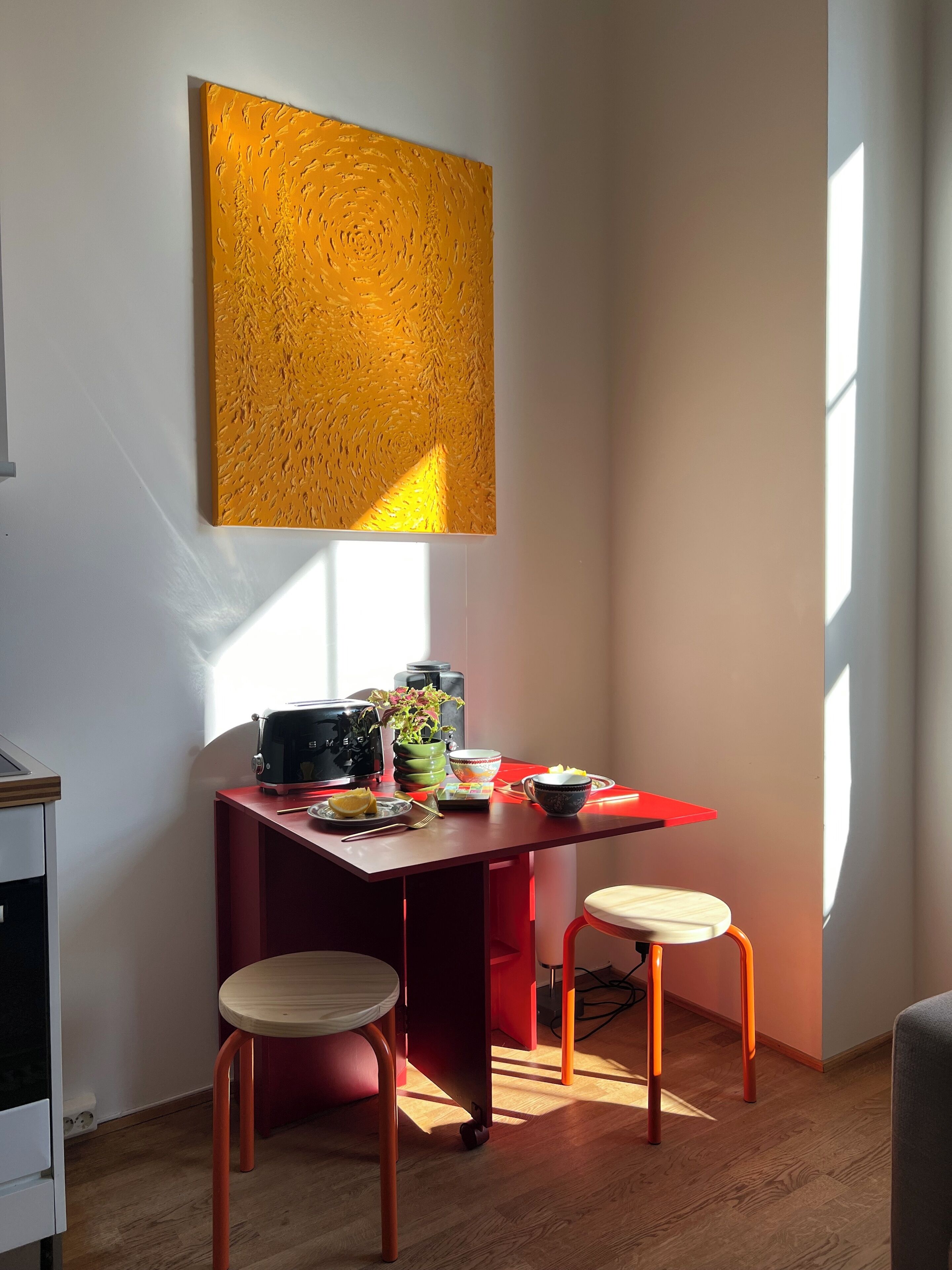 A dining area with a red table, two wooden stools, a black radio, a green plant, and a bowl of fruit.