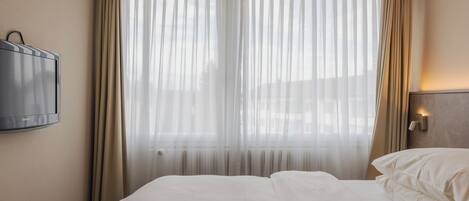 Habitación de hotel con una cama, un televisor montado en la pared y un ventanal con cortinas traslúcidas.