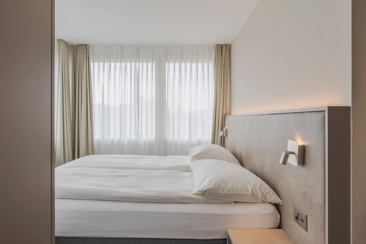 Una cama bien hecha con sábanas blancas, una mesita de noche y una lámpara de pared.