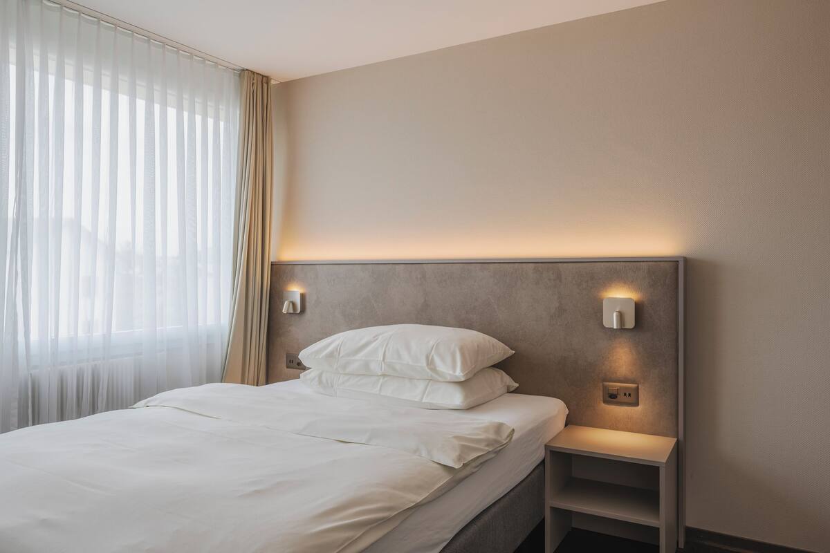 Una cama bien hecha con sábanas blancas, un cabecero con luces integradas y una mesita de noche con toma de corriente.