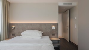 Una habitación de hotel moderna con una cama grande, lámparas de noche, un cabecero y una puerta que da a un pasillo.