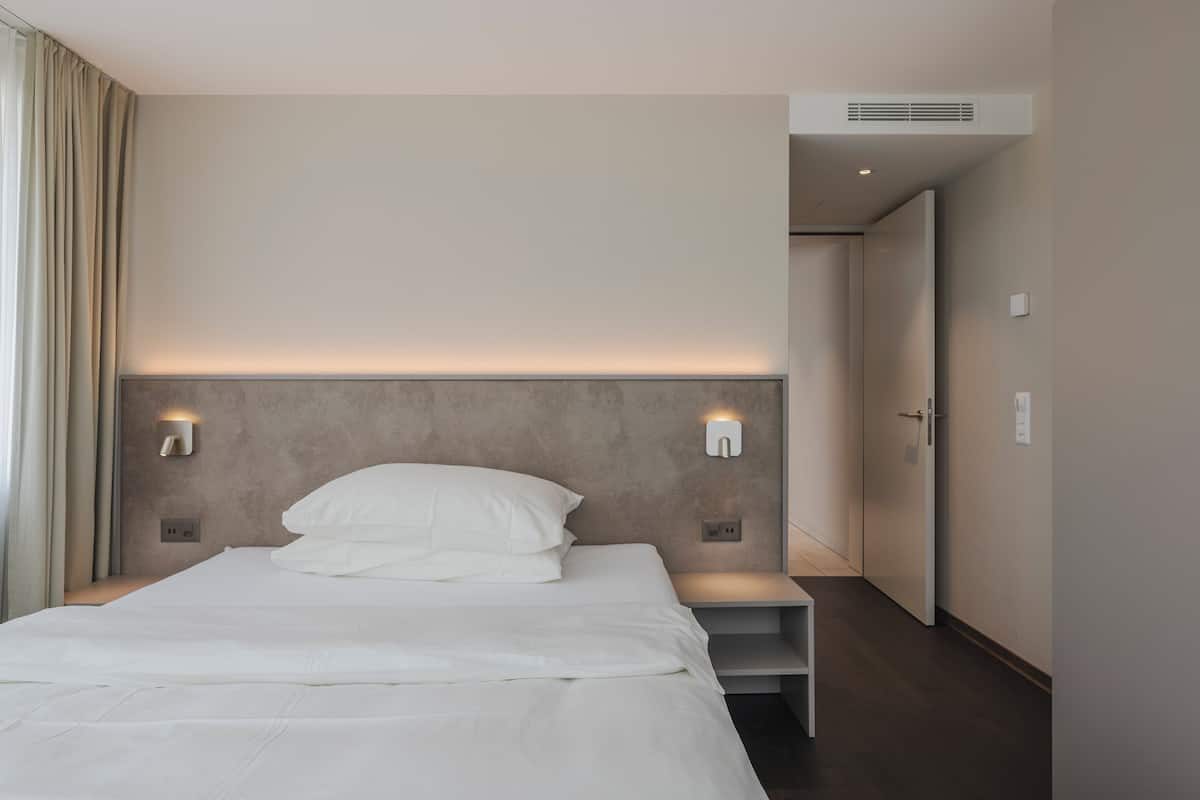 Una habitación de hotel moderna con una cama grande, lámparas de noche, un cabecero y una puerta que da a un pasillo.