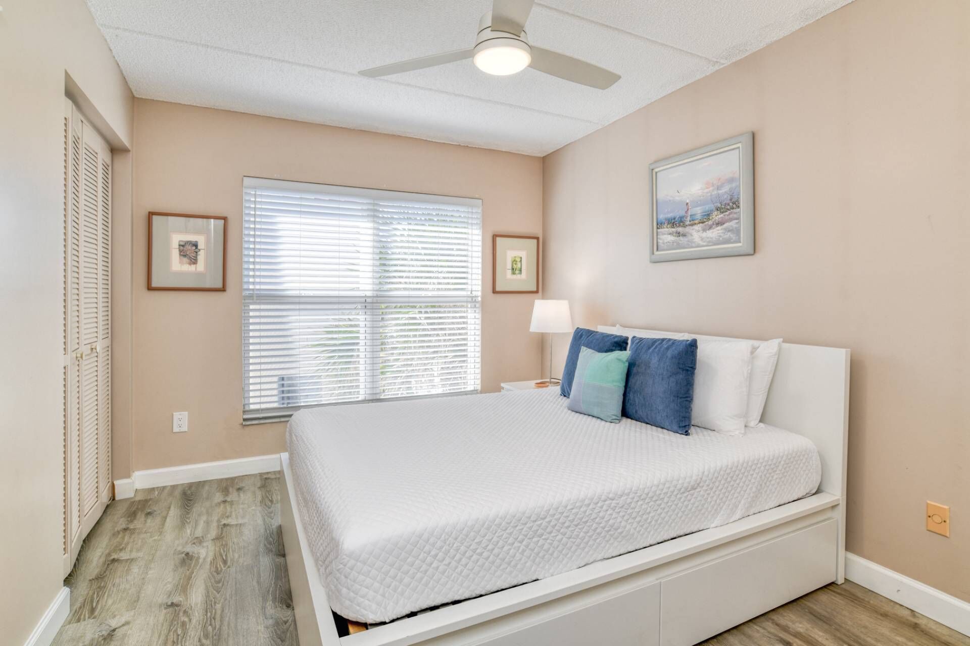Une chambre à coucher avec un lit, un ventilateur de plafond, une fenêtre avec des stores et un tableau accroché au mur.