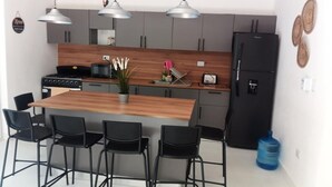 A modern kitchen with a central island, black chairs, pendant lights, and a refrigerator.