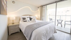 A hotel room with a large bed, a nightstand with a lamp, a balcony with a table and chairs, and a view of the city. - Honolulu Central Waikiki luxury High With Views & Beach (Honolulu)