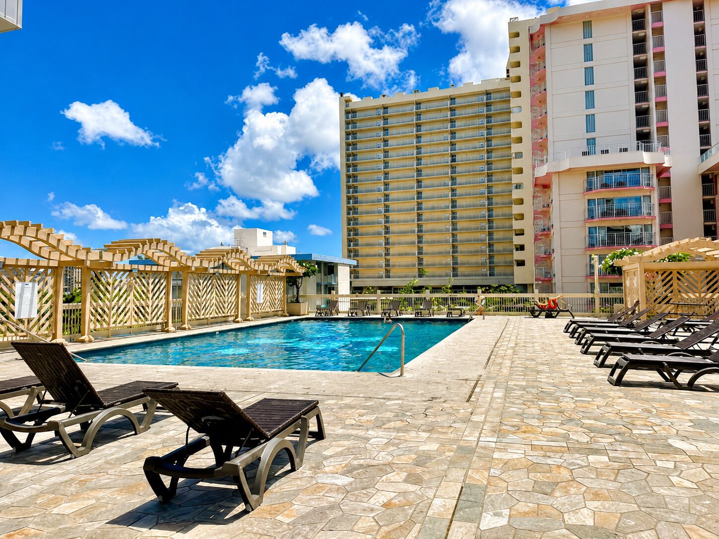 A pool area with lounge chairs, a clear blue sky, and a tall hotel building in the background.