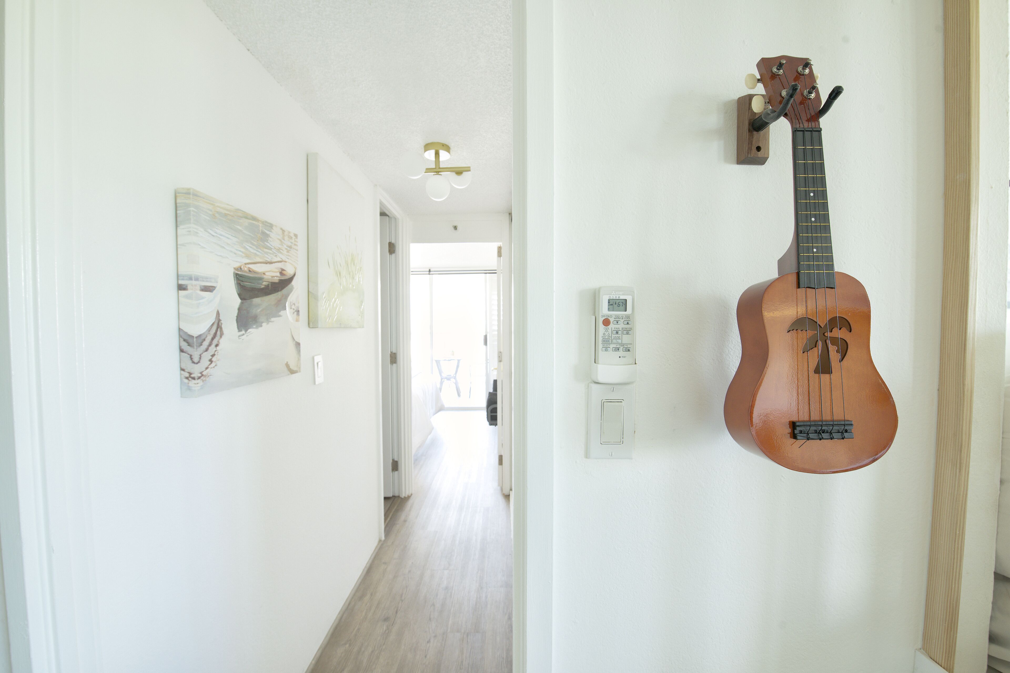 A hallway with a wall-mounted ukulele, a thermostat, and two paintings.