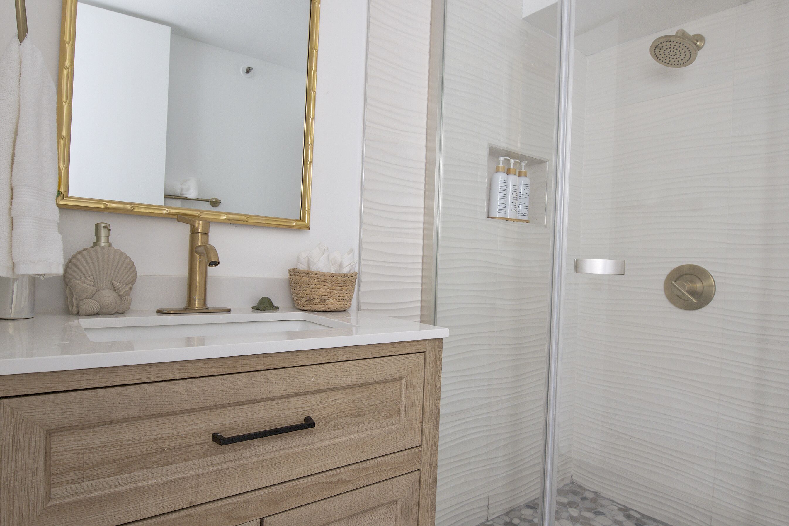 A bathroom with a white vanity, a gold-framed mirror, a wooden drawer, a glass shower enclosure, and a small shelf with toiletries.