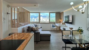 A modern living room with a sofa, ottoman, and a glass dining table. There is a TV mounted on the wall and a window with a city view. - Honolulu Central Waikiki luxury High With Views & Beach (Honolulu)