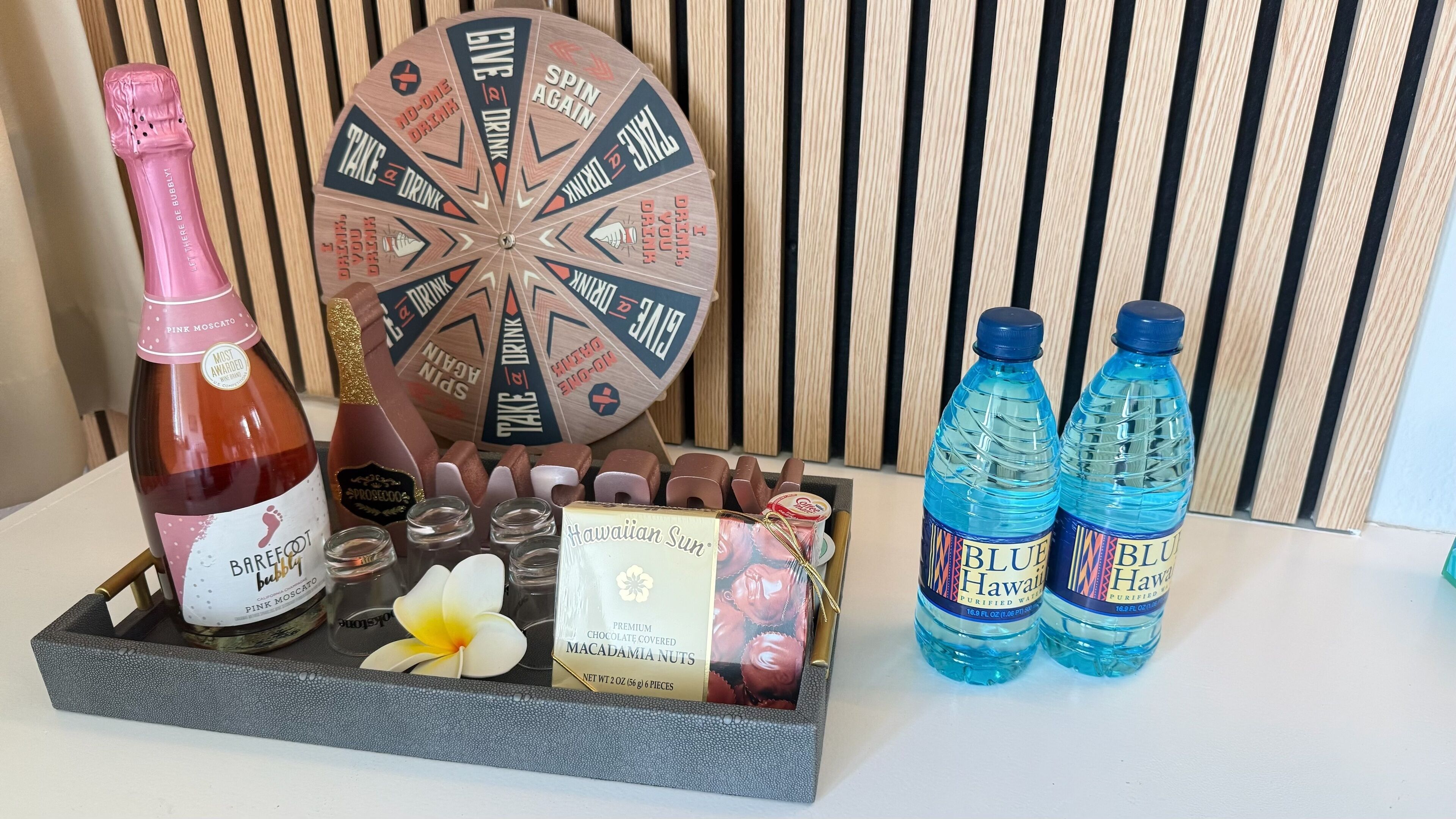 A tray with a bottle of Pink Moscato, a spinning wheel game, chocolate-covered nuts, and two bottles of Blue Hawaiian water.