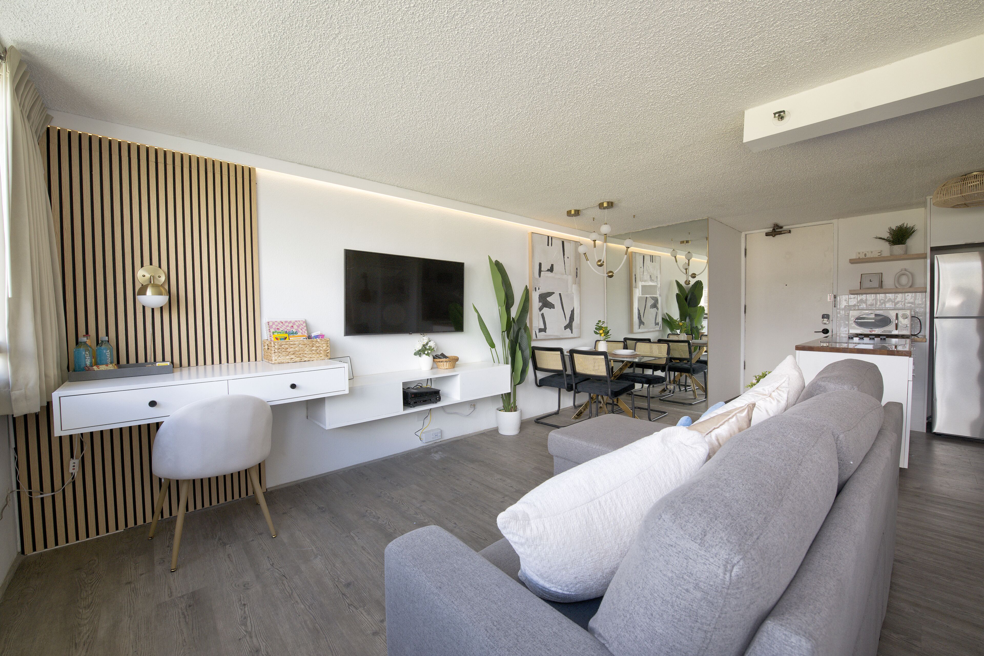 A modern living room with a grey sofa, a white desk, and a flat-screen TV mounted on the wall.
