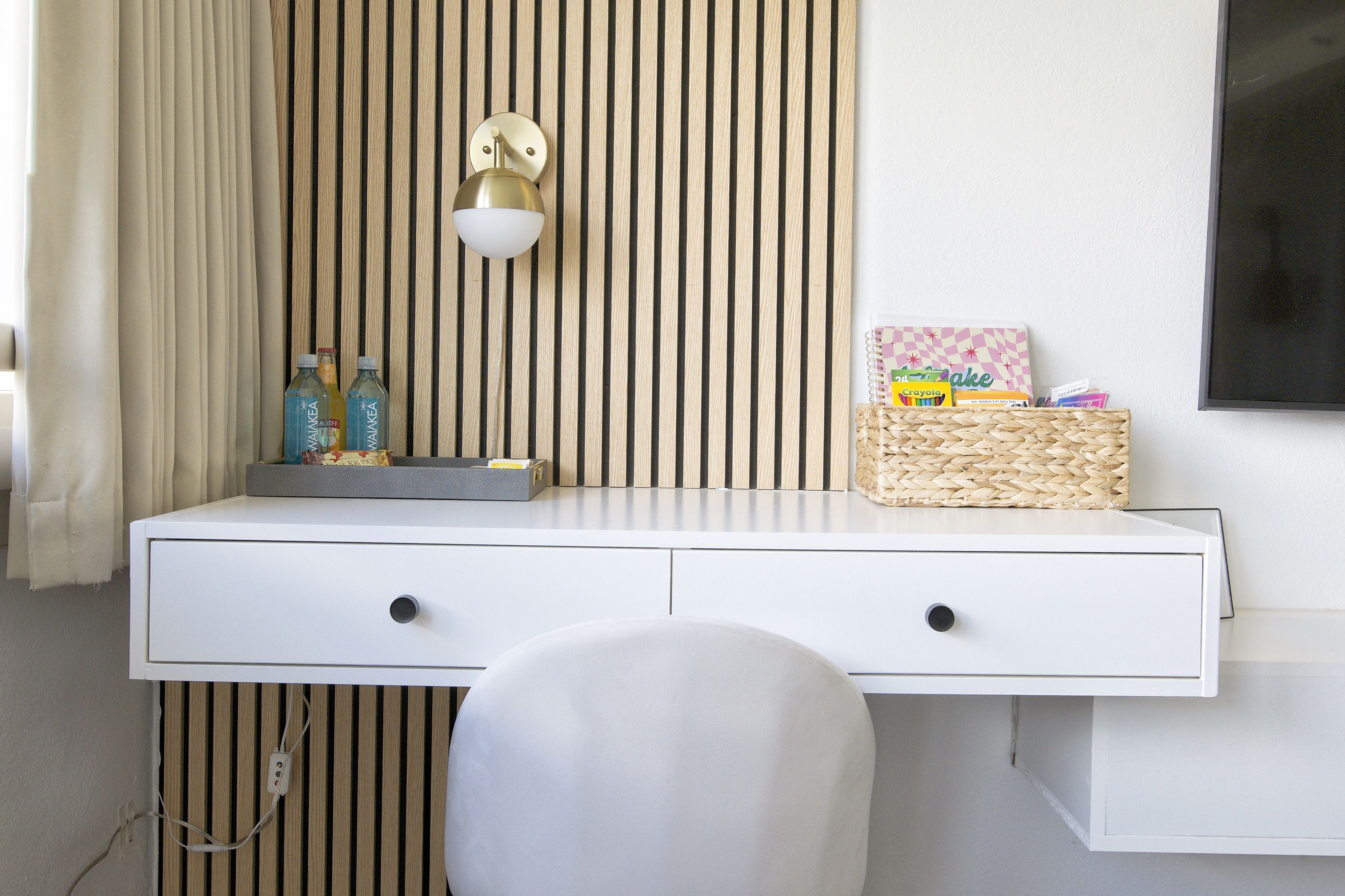 Un bureau blanc avec deux tiroirs, un panier en osier et un luminaire fixé au mur.