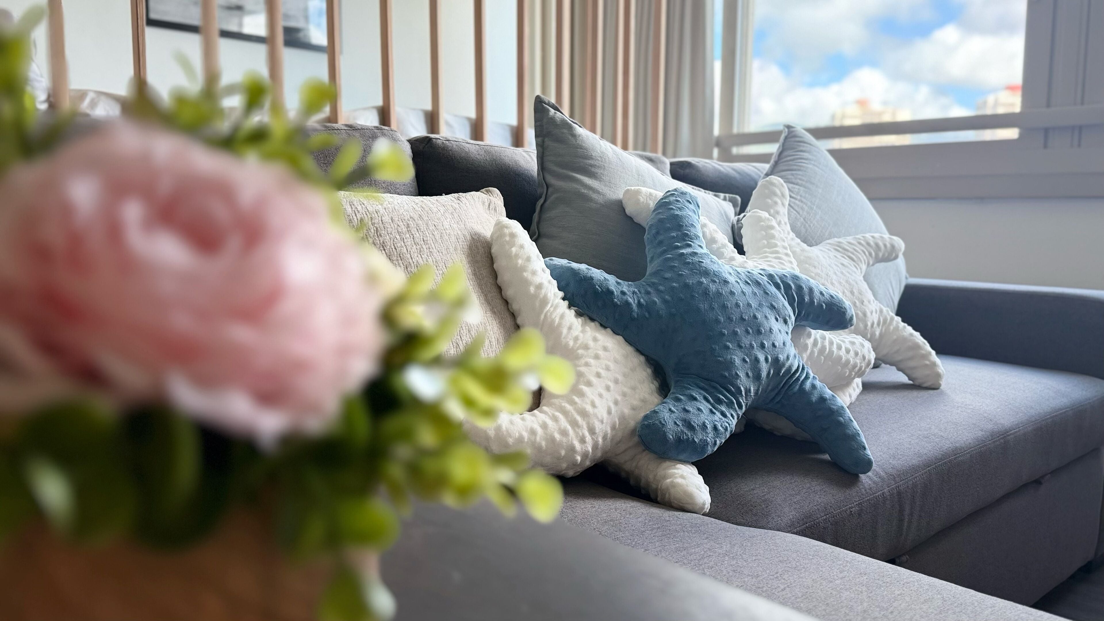 Un sofá gris con varios cojines, incluyendo un cojín azul con forma de estrella de mar, en una habitación con una ventana y una planta en primer plano.