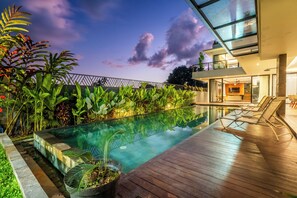 Ein moderner Poolbereich im Freien mit Holzterrasse, Liegestühlen und üppiger tropischer Bepflanzung.
