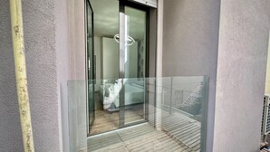 Una habitación de hotel moderna con balcón de cristal, una cama y un baño con ducha y lámpara colgante.
