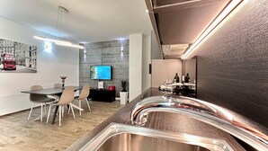 Una cocina moderna con un lavamanos de acero inoxidable, una zona de comedor con mesa y sillas, un televisor de pantalla plana montado en una pared con textura, y una planta pequeña en la encimera.