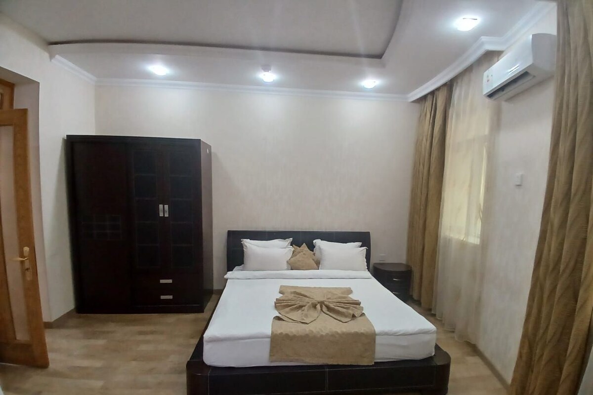 A neatly made bed with a decorative headboard, a wardrobe, and an air conditioning unit in a well-lit room.