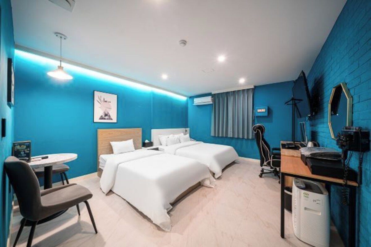 A modern hotel room with two beds, a desk, a chair, and a small table.