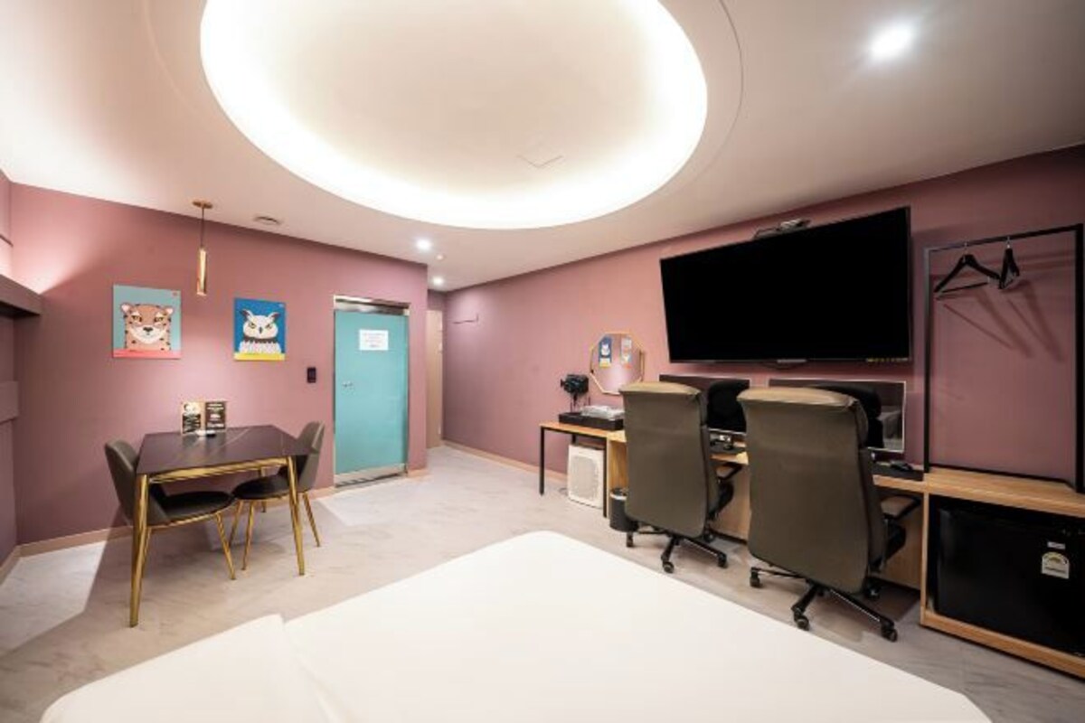 A hotel room with a round ceiling light, a TV, a desk with two chairs, a dining table with chairs, and a wardrobe.