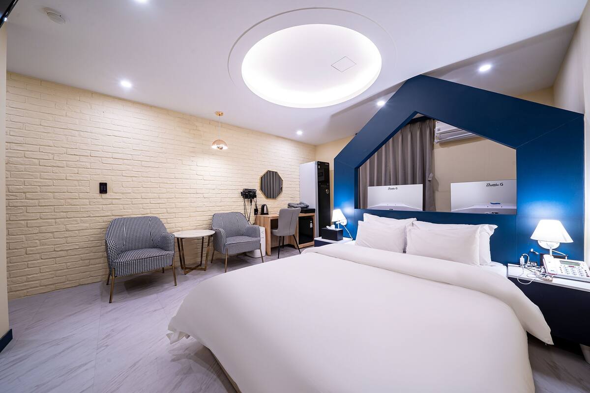 A modern hotel room with a large bed, a desk, a chair, a small table, a mirror, and a phone on the nightstand.
