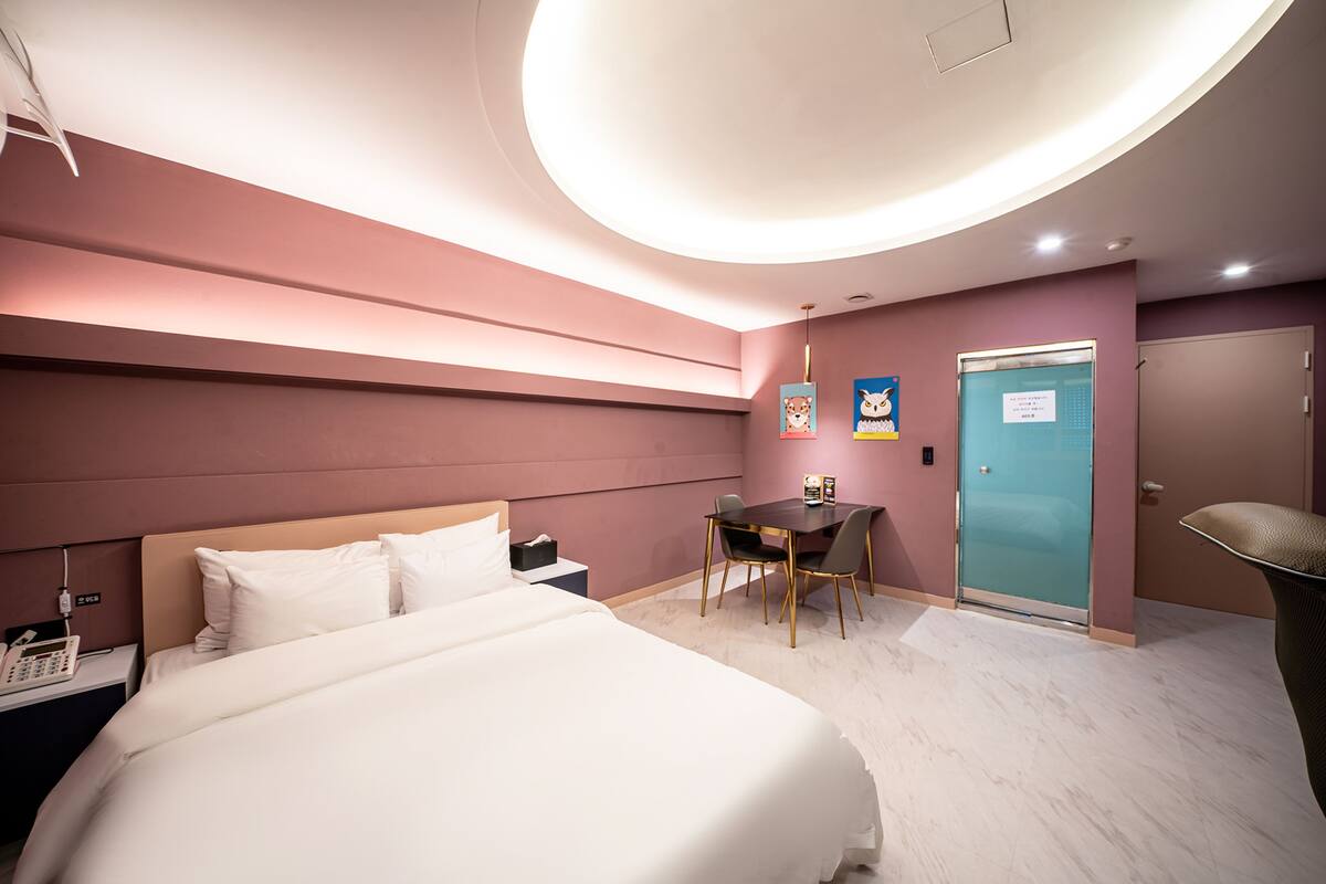 A hotel room with a large bed, a desk with chairs, a door, and a wall-mounted light fixture.