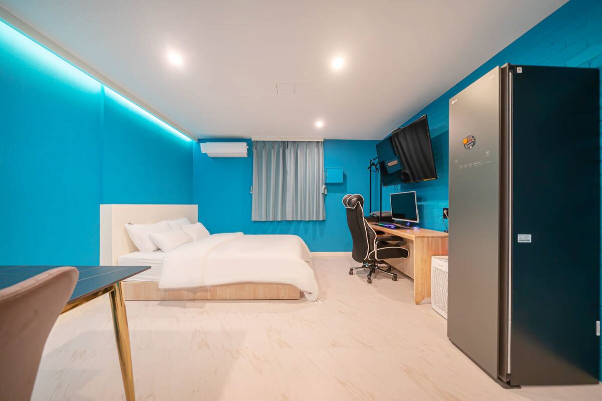 A modern hotel room with a bed, desk, chair, and a large refrigerator.