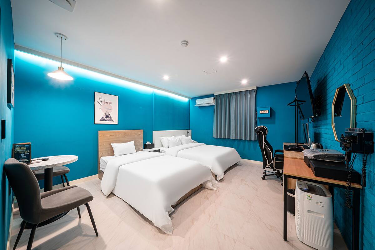 A modern hotel room with two beds, a desk with a computer, a chair, and a small table.