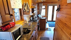A kitchen with wooden cabinets, a stove, and a microwave. There is a window with curtains and a rug on the floor. - Riverside chalet in Aviemore, short walk to centre (Aviemore)