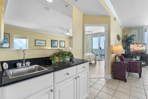 A modern kitchen with a black countertop, white cabinets, and a sink. There is a ceiling fan, a plant on the counter, and a view of the living area with a chair and a lamp. - San Carlos 1908 (Gulf Shores)