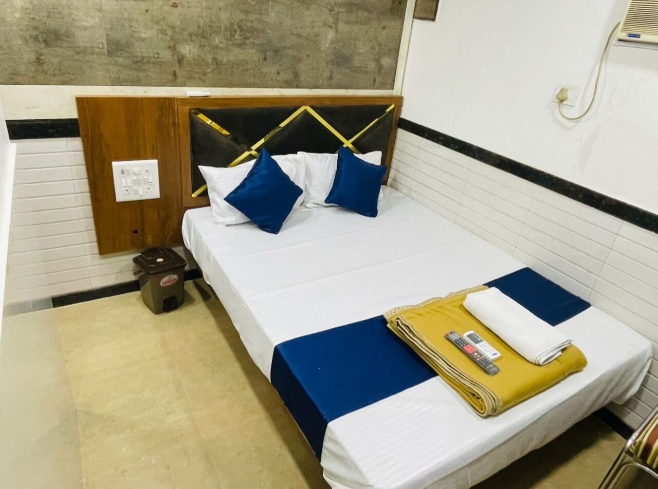 A single bed with a wooden headboard, white bedding, and blue pillows. A small trash bin is visible to the left of the bed.