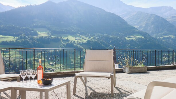 A terrace with a table, two chairs, a bottle, and glasses. A mountain view is visible in the background.