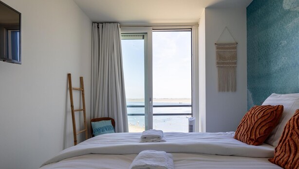 A hotel room with a bed, a view of the beach, a ladder, and a wall-mounted macrame piece. - Lux app Vista M - Havenweg 8- 40 (Sint-Annaland)