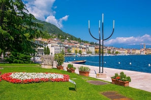 A waterfront promenade with a row of colorful buildings, a boat docked by the water, and a decorative metal structure. - Ricomilla Holiday - Ulivo Innamorato (Roe Volciano)
