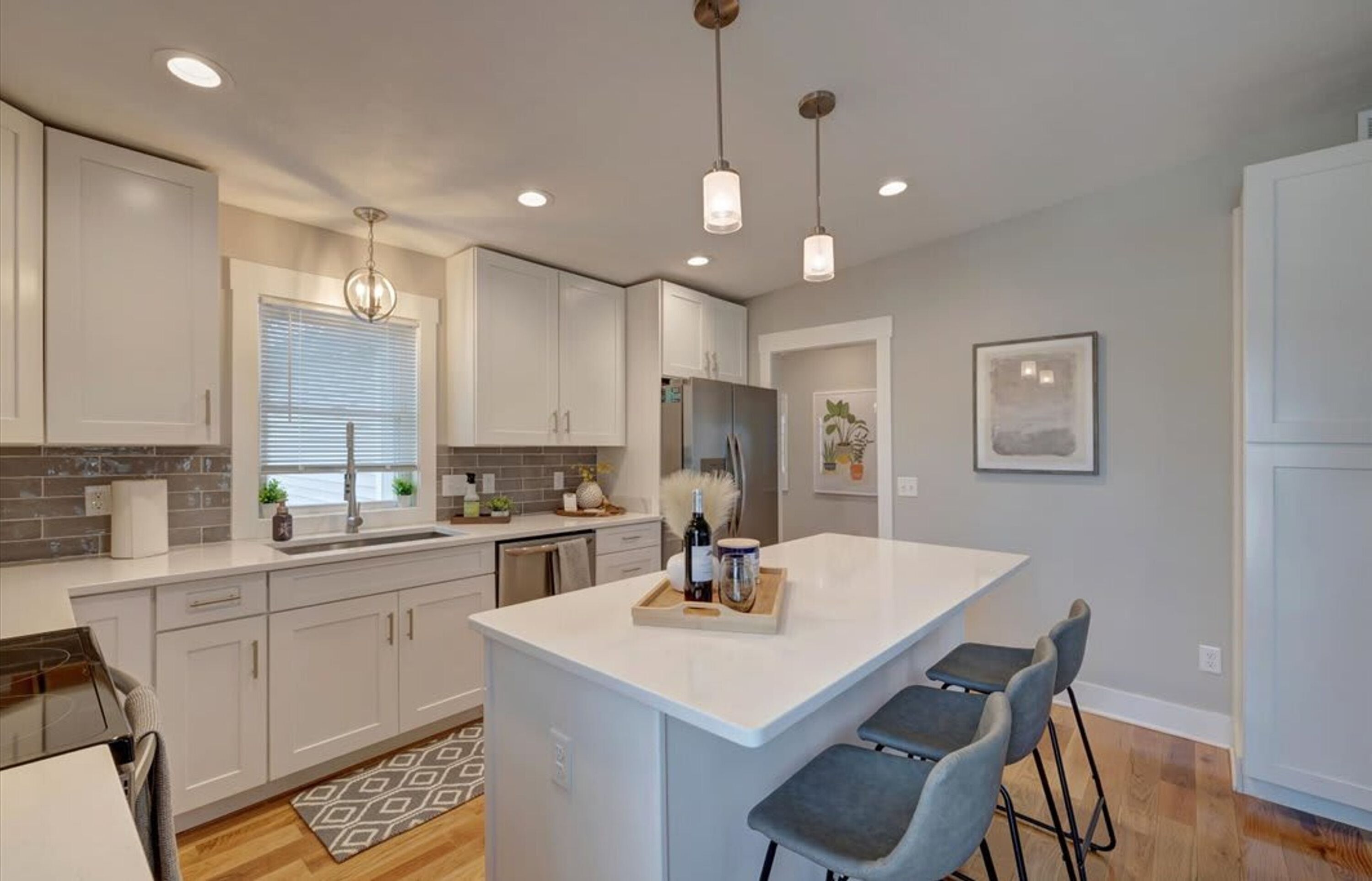 A modern kitchen with white cabinetry, a central island with bar stools, pendant lighting, and a window with blinds.