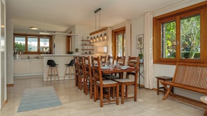 A spacious dining area with a wooden table and chairs, a kitchen in the background, and large windows with a view of greenery. - Villa Azur on Lake Neuchâtel (La Grande Béroche)