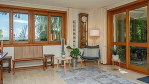 A spacious living room with a wooden bench, a chair, a table, a clock, and large windows offering a view of the outdoors. - Villa Azur on Lake Neuchâtel (La Grande Béroche)