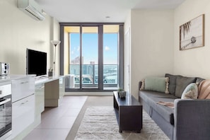 A modern apartment with a kitchen, dining area, and living room. It features a large window with a city view, a flat-screen TV, and a sofa with cushions.